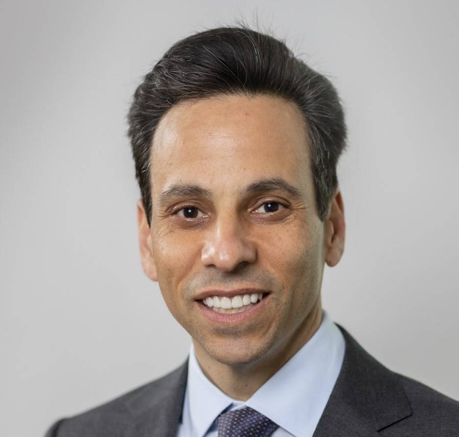 A photo of David Senior wearing a dark gray suit, white collared shirt, and dark blue tie with a diamond pattern